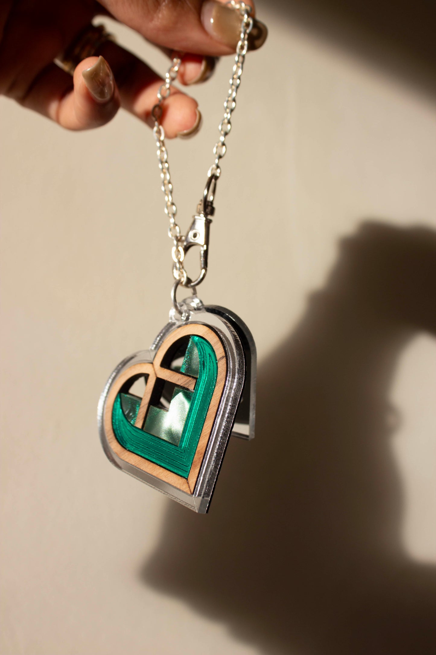 Heart-shaped acrylic bag charm with green and silver accents on a chain, held by a hand against a beige background.