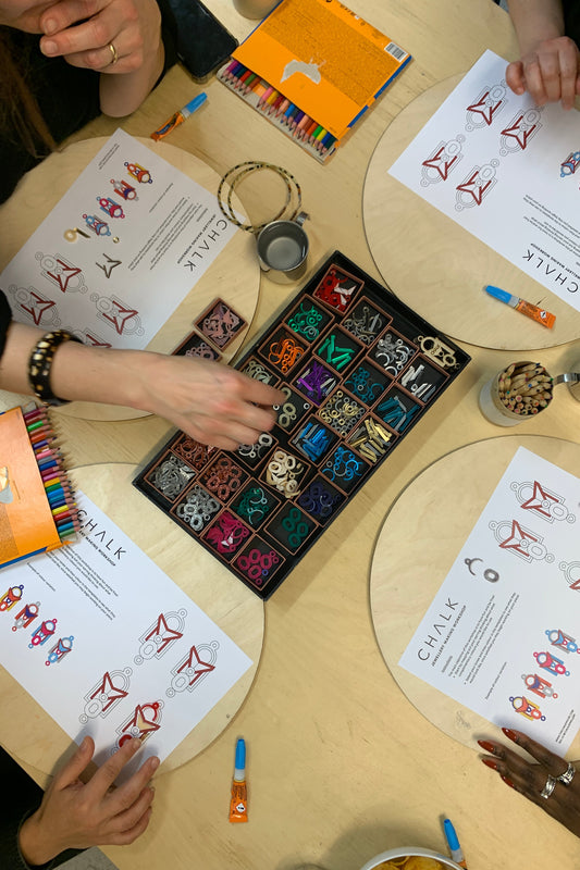 Table with workshop supplies, including a box of colored acrylic pieces and worksheets, with people working.