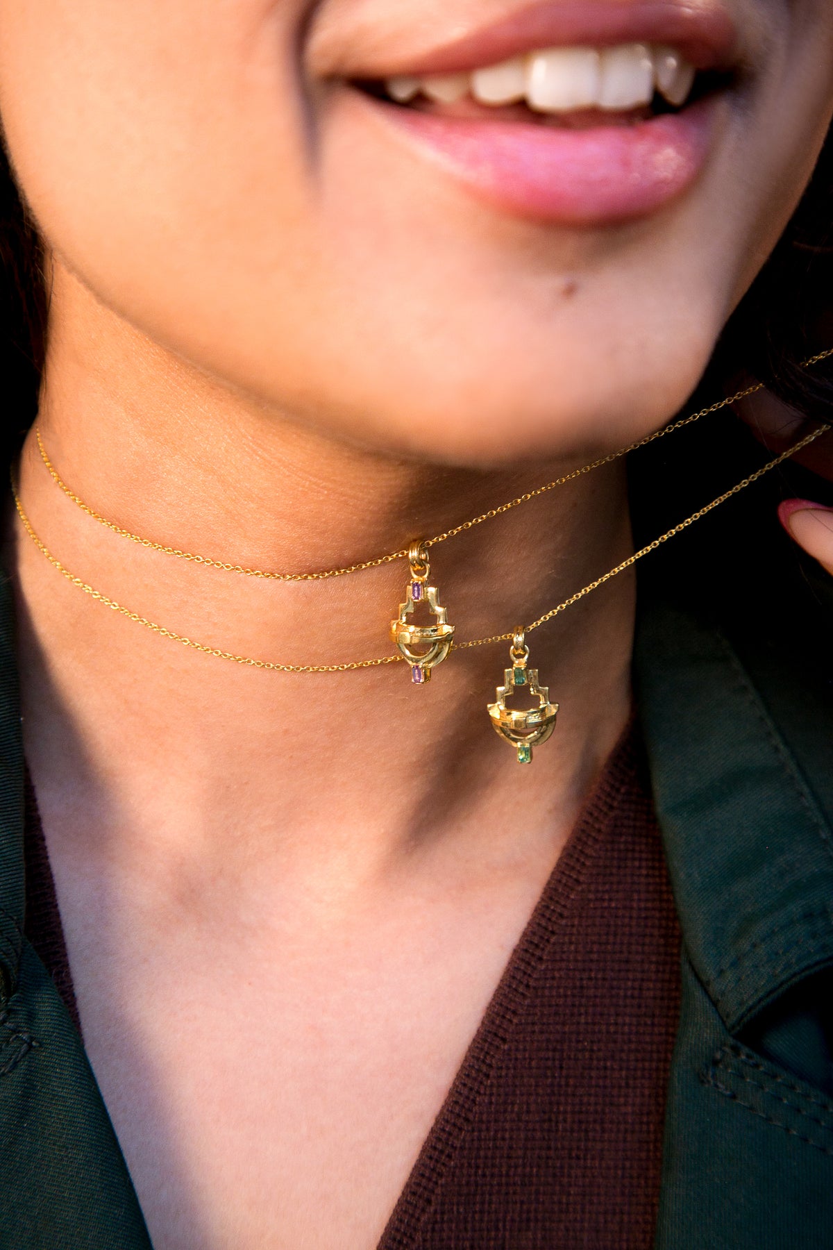 close up of a woman wearing gold geometric necklaces with green tourmaline and purple amethyst gemstones against a beige background