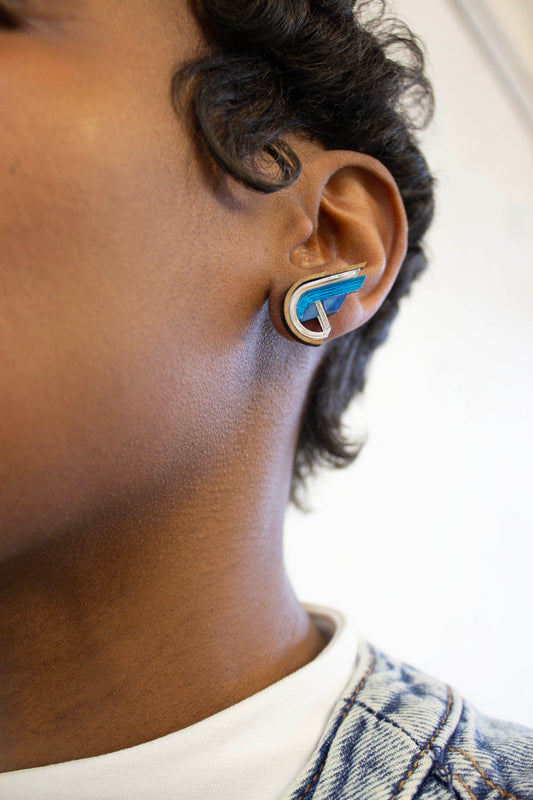 Close-up of an ear wearing a blue and silver half heart shaped earring with a white background. Made from acrylic, walnut wood and plywood