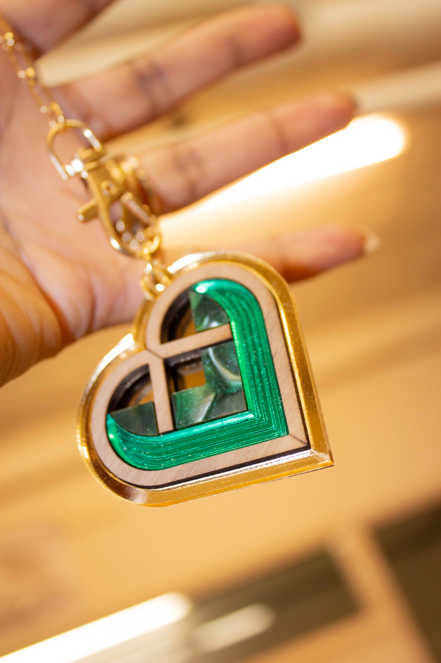 Heart-shaped bag charm with green and gold design held by a hand on a reflective surface. Laser cut charm made from plywood, walnut wood and acrylic