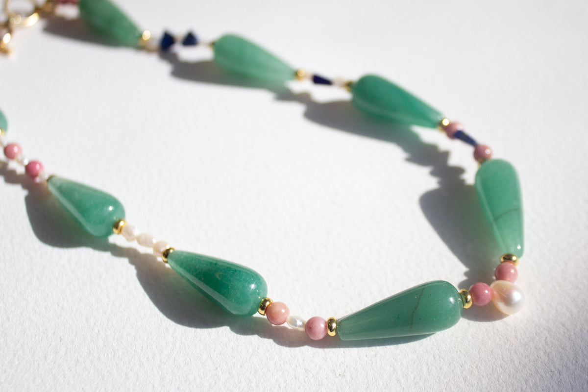 A beaded necklace featuring large green teardrop-shaped beads, small pink round beads, white pearl-like beads, and tiny gold spacer rings, arranged on a white background.