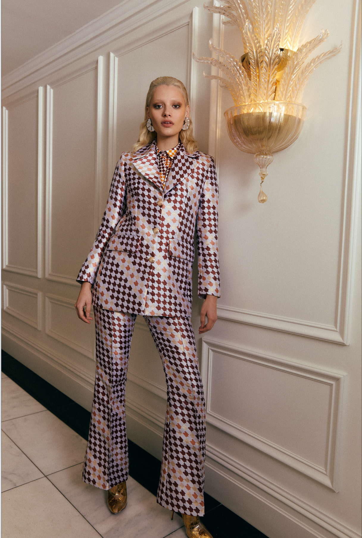 Woman wearing silver earrings with gold accents, standing in an elegant room with decorative wall lights. Laser cut earrings made from acrylic, walnut wood and plywood.